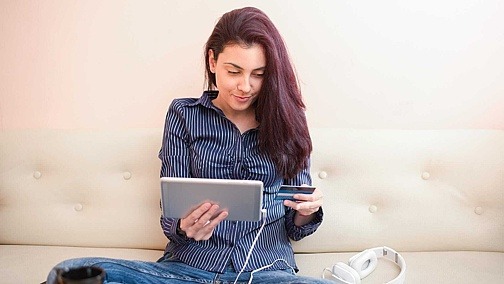 Virtuelle Währung: Wir fair sind In-Game-Käufe? Junge Frau sitzt mit dem Tablet auf der Couch und vergleicht ihre Kreditkarteninformationen.